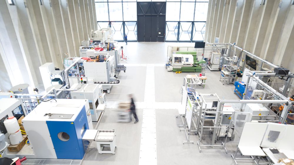 Auf dem Bild ist das Innere der ETA-Fabrik zu sehen. In einem hellen Fabrikraum arbeiten menschen an verschiedenen Produktionsanlagen.